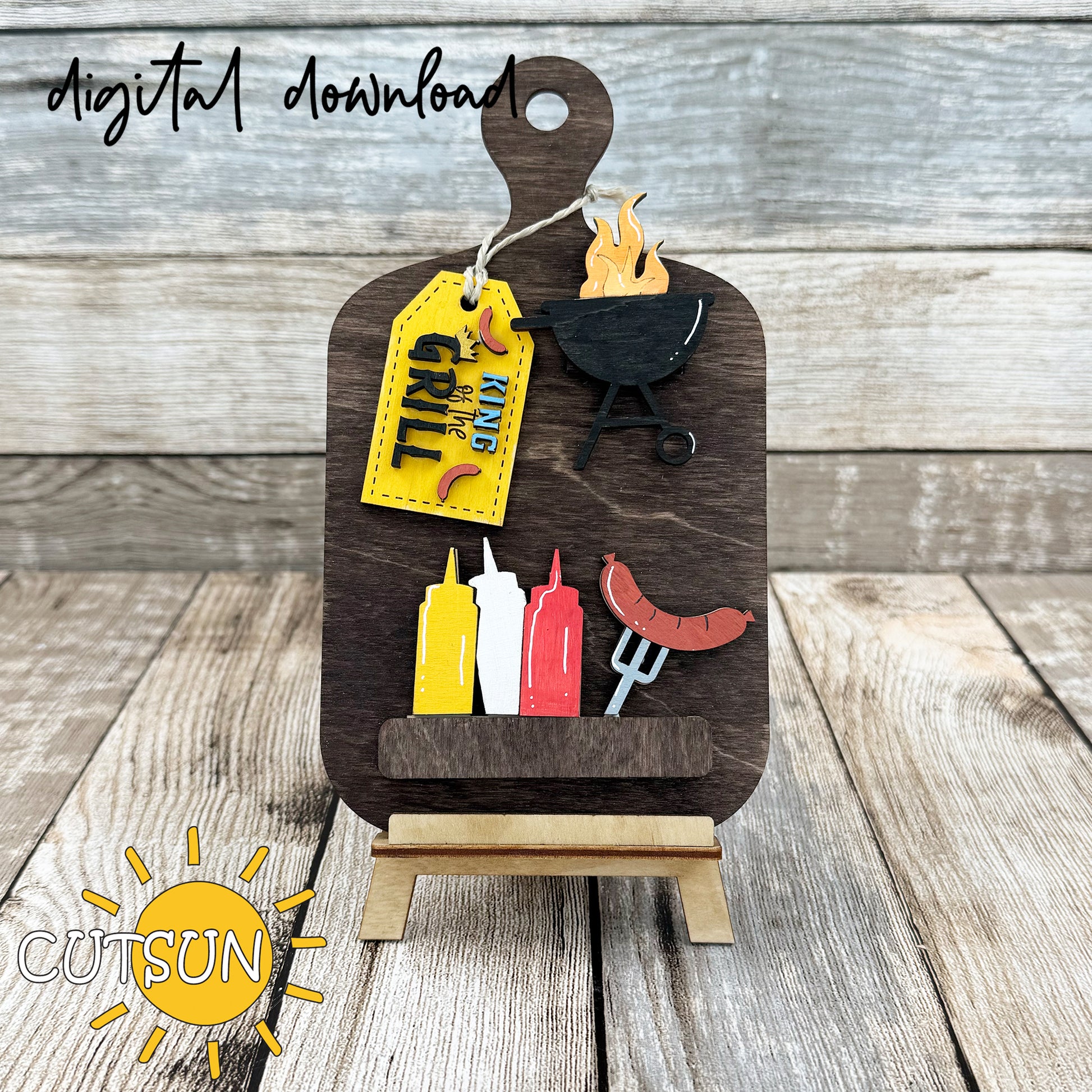 A digital representation of a laser-cut wooden cutting board decor with interchangeable elements including a barbecue, condiment bottles, a fork with a sausage, and a 'King of the Grill' tag.