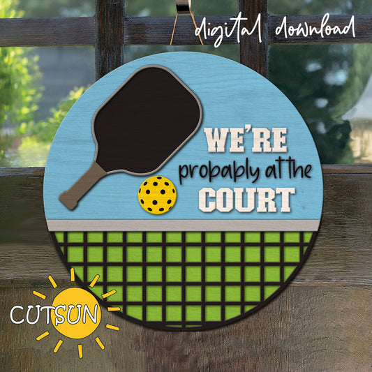 Decorative sign with pickleball paddle and ball design, text 'WE'RE probably at the COURT', and brand logo on a wooden background.