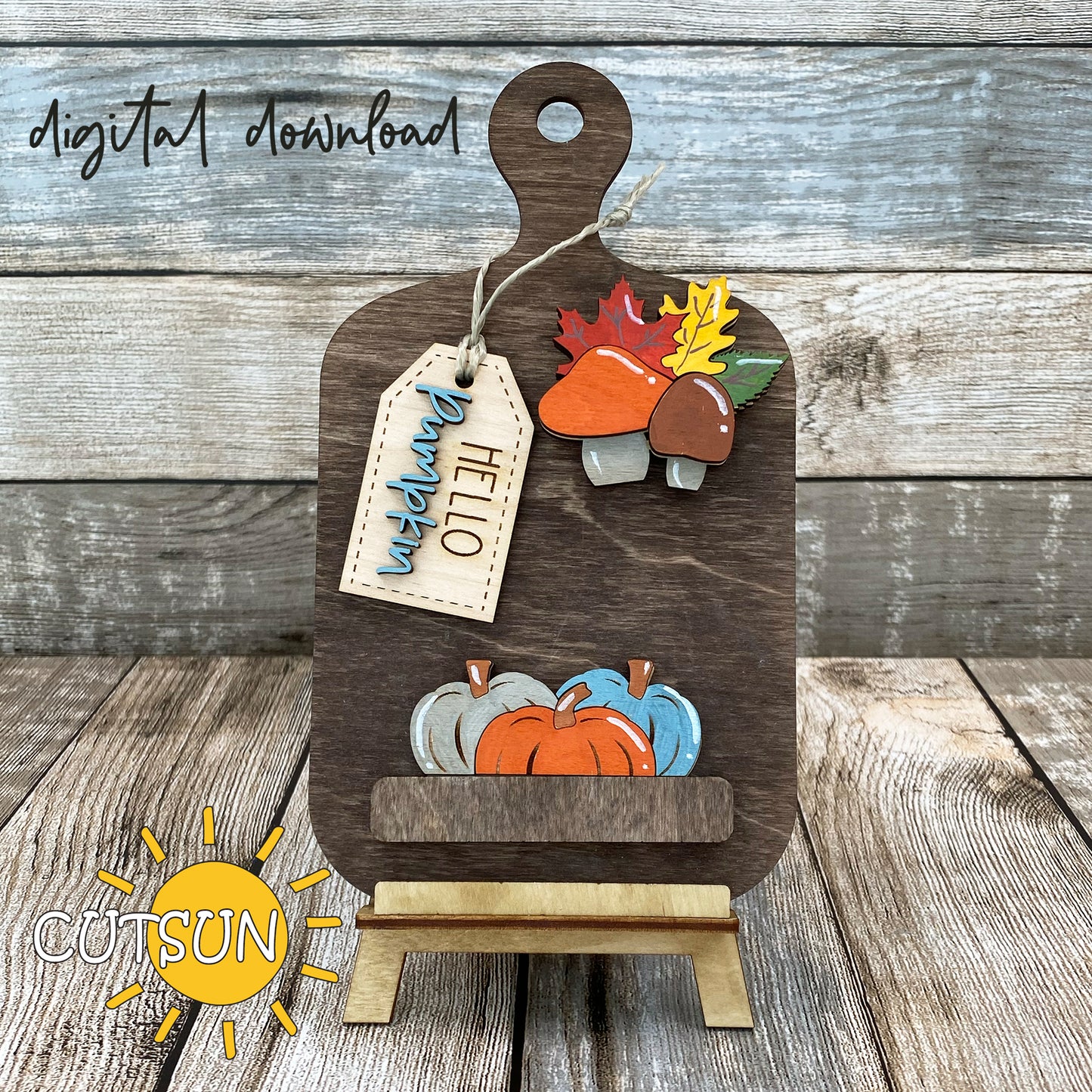 A digital download of a laser cut file for a cutting board decoration featuring fall leaves, mushrooms, pumpkins, and the text 'Hello Pumpkin'. The image shows the decoration in a wooden style with a tag, on a wooden background.