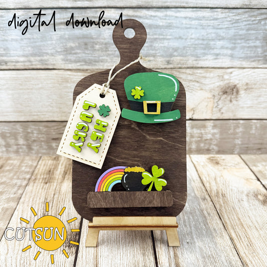 A digital illustration of a wooden cutting board decoration with a St Patrick's day theme, featuring a leprechaun's hat, a four leaf clover, a rainbow, a pot of gold and shamrock, and a tag with the words 'Hey Lucky'. The text 'DIGITAL DOWNLOAD' indicates the product is available as a digital file for cutting machines like Glowforge and Muse.
