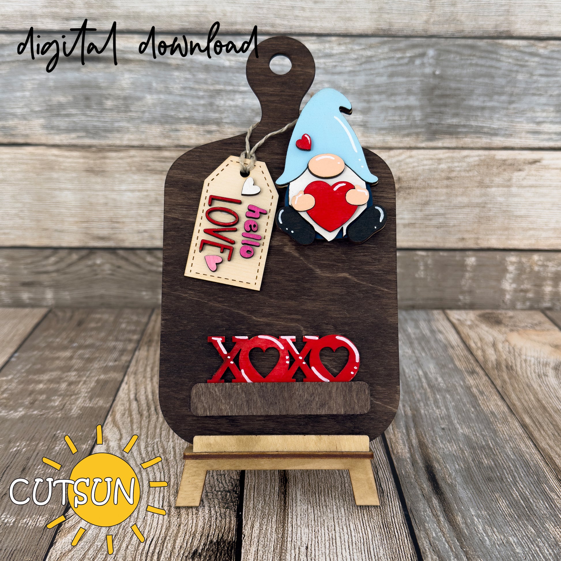 A digital download of a laser cut file featuring an interchangeable cutting board decor with a Valentine's day theme. The image shows a gnome holding a heart, large XOXO letters, and a tag with the words 'Hello Love'.