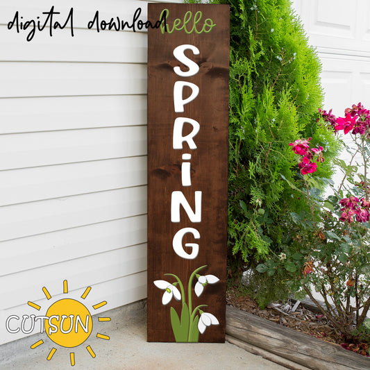 Wooden sign with 'hello spring' text and floral design against a white wall with plants.