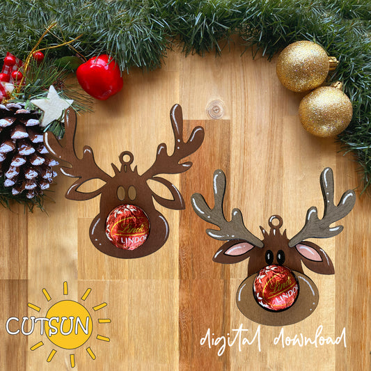 Two Christmas reindeer-shaped ornaments displayed on a wooden surface with holiday decorations around the edges, including pine branches, ornaments, and a pinecone. The ornaments are dark brown with antlers and pink inner ears, each holding a wrapped candy.