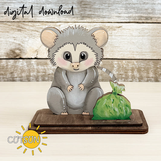 Layered wooden opossum shelf sitter with large eyes sitting next to a green trash bag on a rectangular base, designed as interchangeable laser cut decor