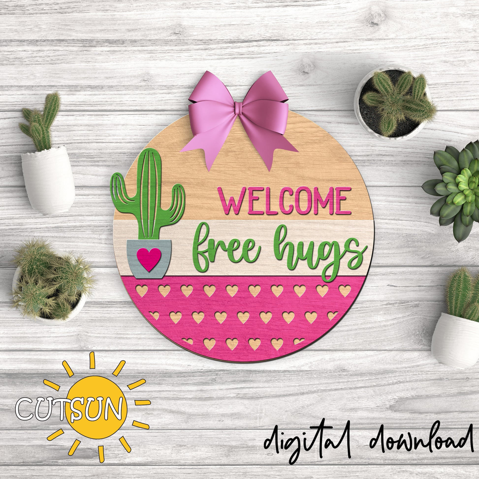Decorative sign with cactus, pink bow, and 'Welcome free hugs' text on a wooden background with plants.